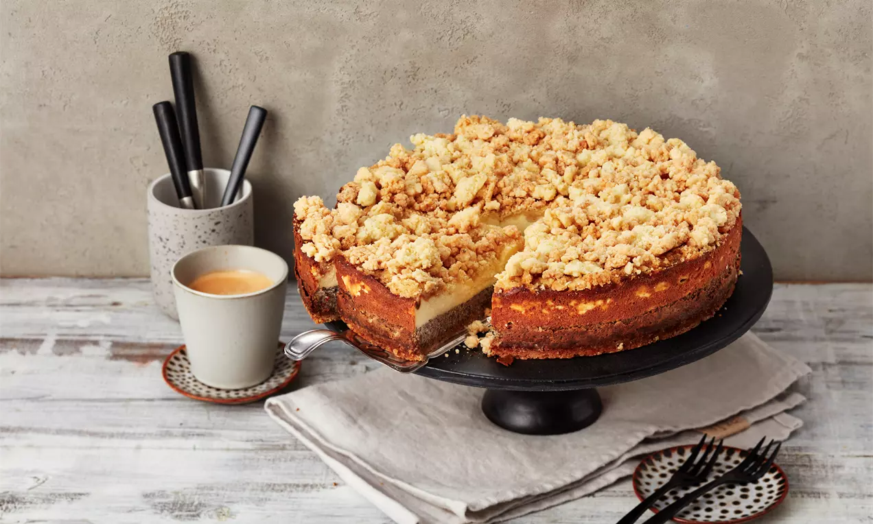 Mohn-Käsekuchen mit goldbraunen Streuseln, cremiger Füllung und Mohnschicht auf schwarzer Tortenplatte, umgeben von Kaffee und Besteck