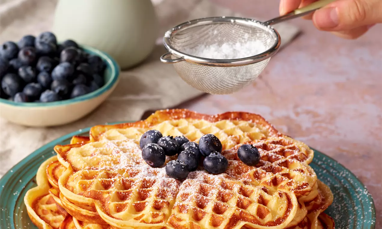 Goldbraune Waffeln in Herzform übereinander gestapelt, mit Puderzucker bestreut und frischen Blaubeeren garniert auf einem grünen Teller
