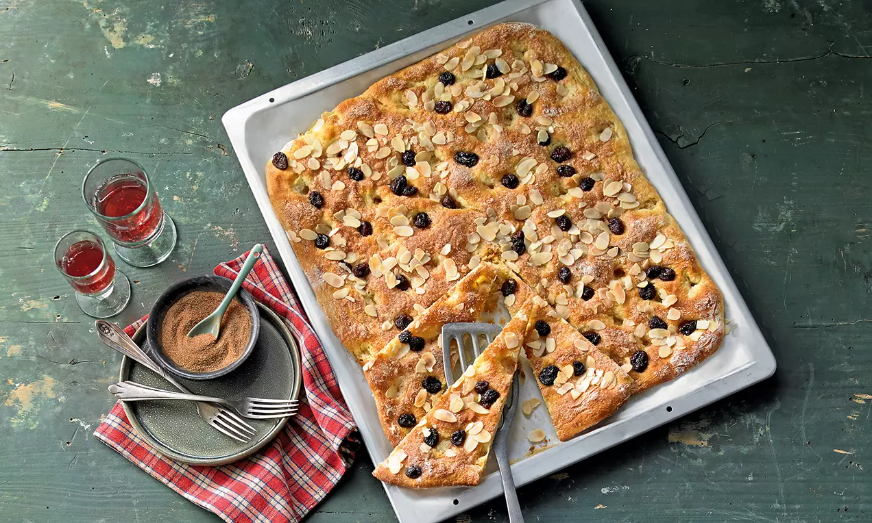 Goldbrauner Kartoffelkuchen mit knusprigem Teigrand, Mandelsplittern und Rosinen auf einem silbernen Blech, daneben Gläser und Geschirr