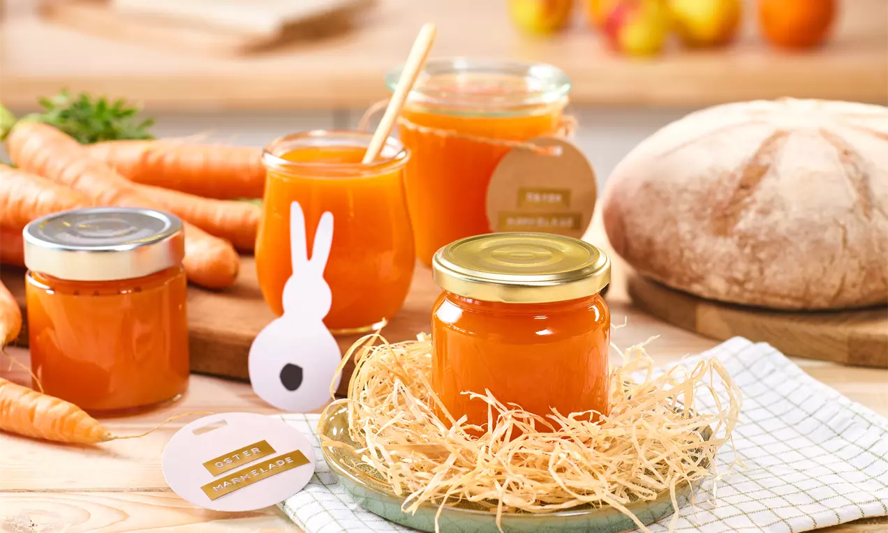 Glas mit orangefarbener Oster-Marmelade in einem Nest aus Holzwolle, umgeben von weiteren Marmeladengläsern, frischen Karotten, einem Laib Brot und österlicher Deko in hellem Küchen-Setting.