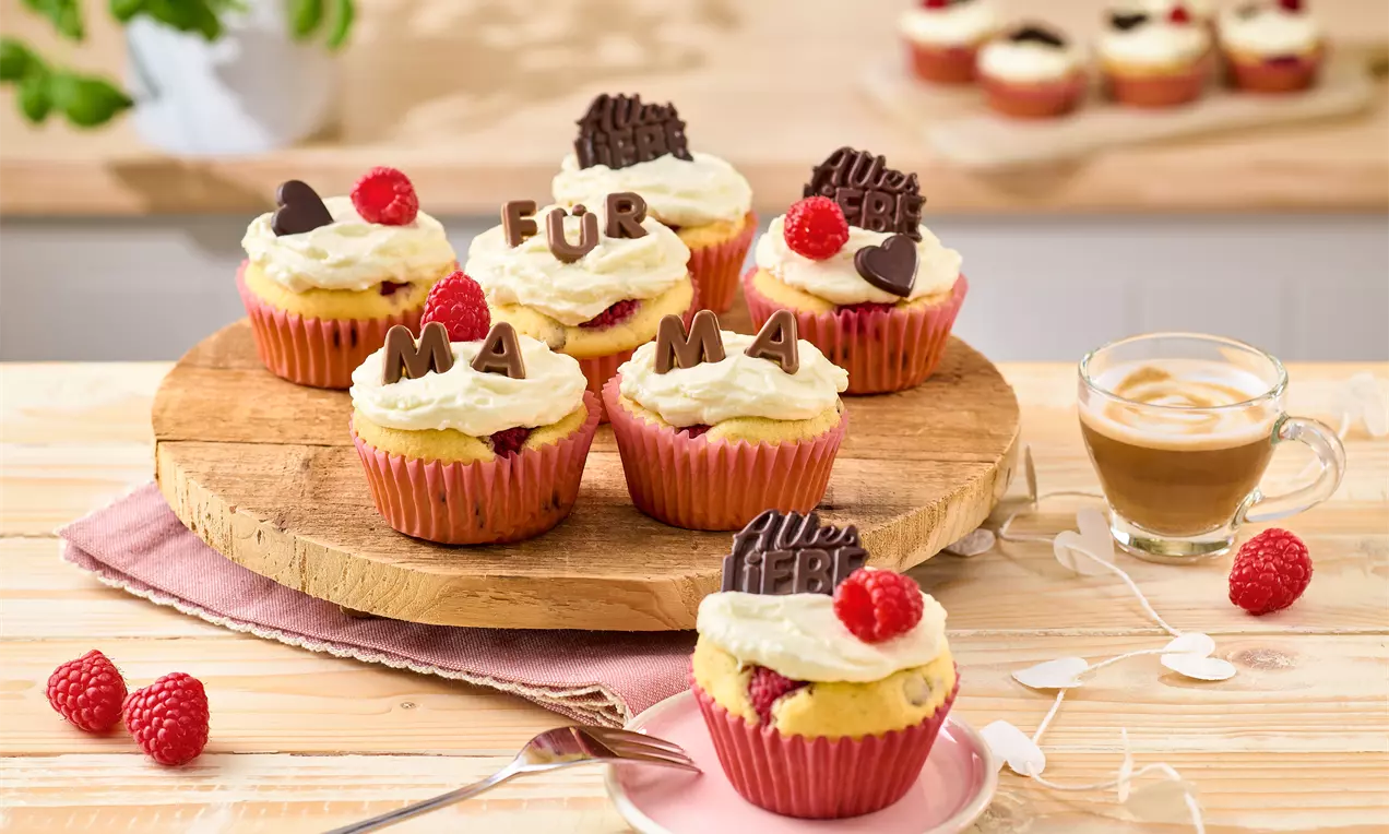 Muffins mit heller Creme, roten Himbeeren und Schoko-Deko auf rosa Förmchen, arrangiert auf rustikalem Holzbrett mit Kaffeetasse