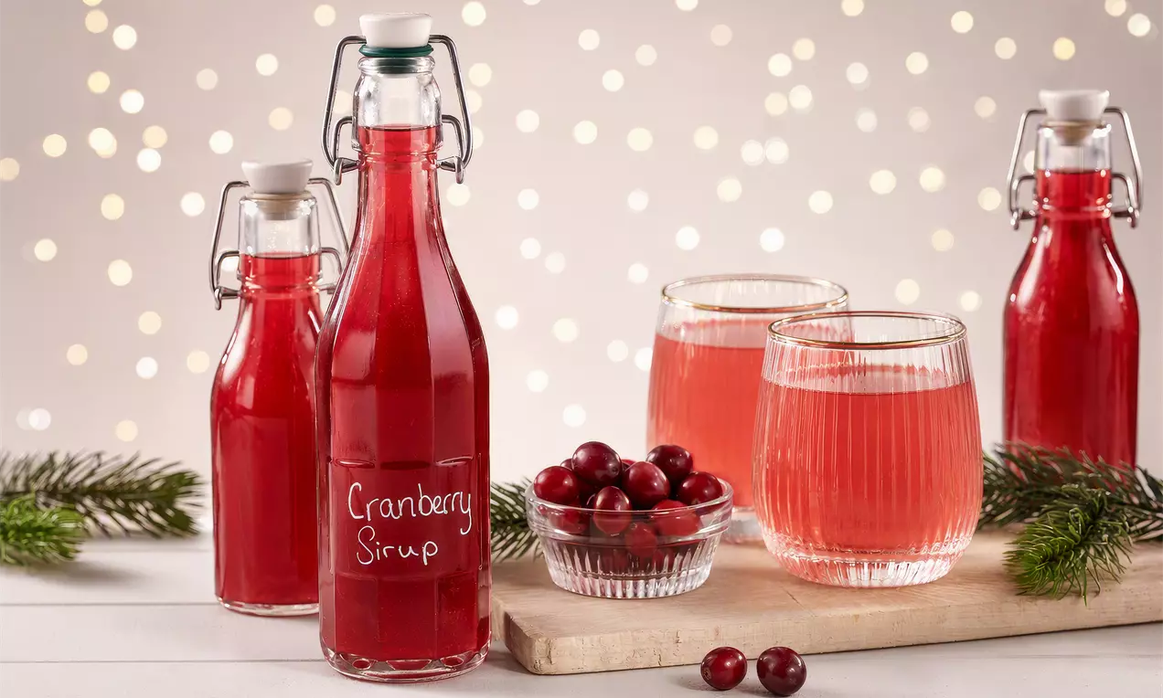 Cranberrysirup in Flaschen und Gläsern mit leuchtend roter Farbe auf hellem Holzbrett, dekoriert mit Cranberries und Tannenzweigen, AI-labeled image