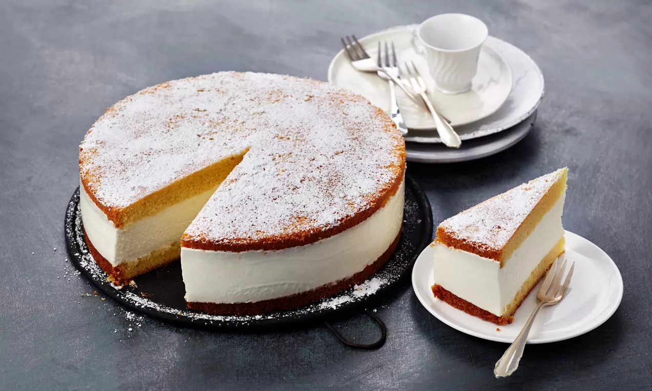 Käse-Sahne-Torte mit goldbraunem Boden und hellem Sahnebelag, bestäubt mit Puderzucker, auf schwarzer Platte und weißem Teller angerichtet