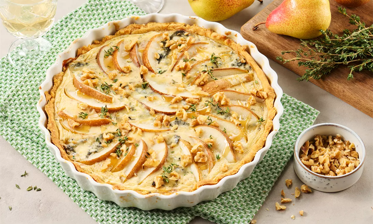 Goldbraune Quiche in weißer Tarte-Form, belegt mit Birnenscheiben, zerbröselten Walnüssen, Gorgonzola und frischem Thymian.