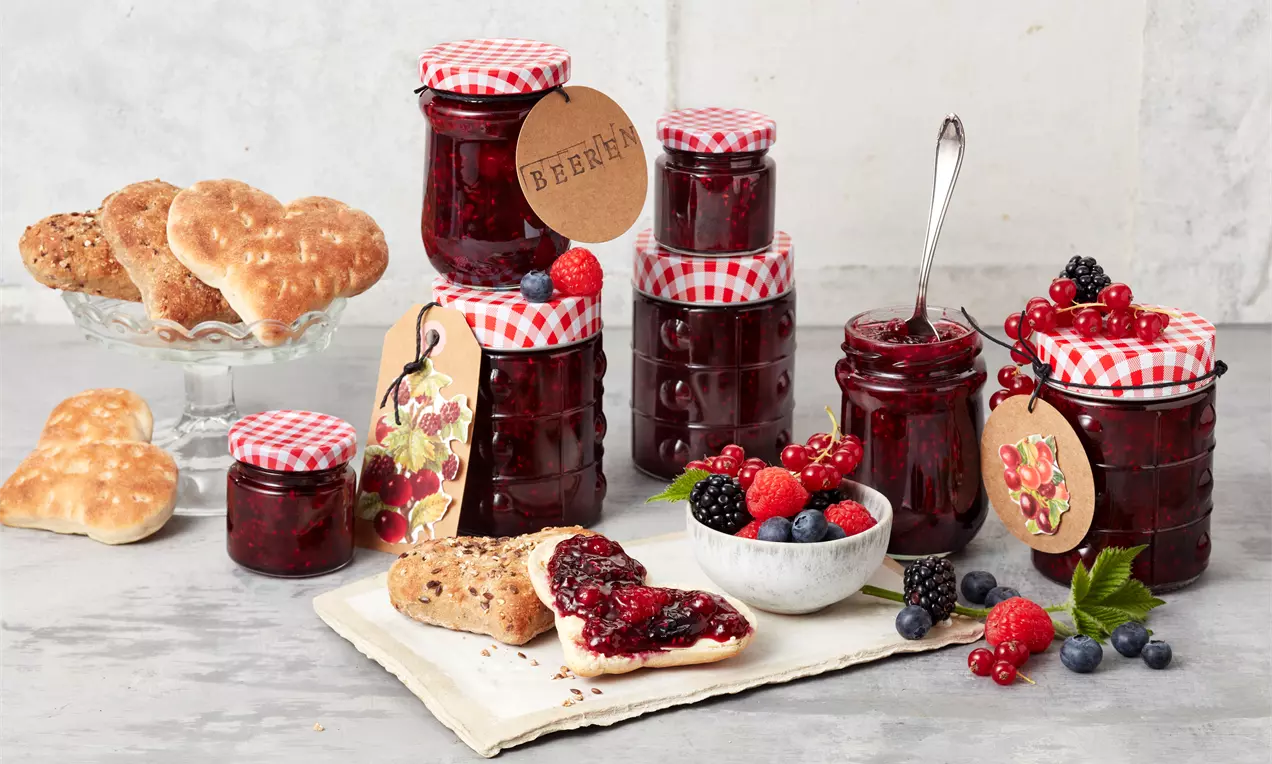 Gläser mit dunkelroter Beerenmarmelade und rot-weiß karierten Deckeln, dekoriert mit Beeren und Herzgebäck auf einer hellen Platte
