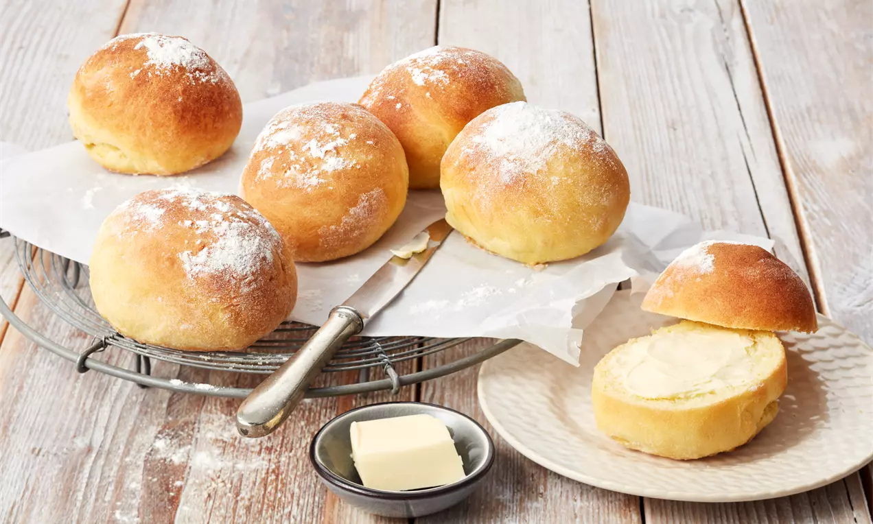 Brötchen ohne Hefe auf rustikalem Holzbrett, goldbraune, knusprige Krusten, weiches Inneres sichtbar, dazu Butter und frische Kräuter