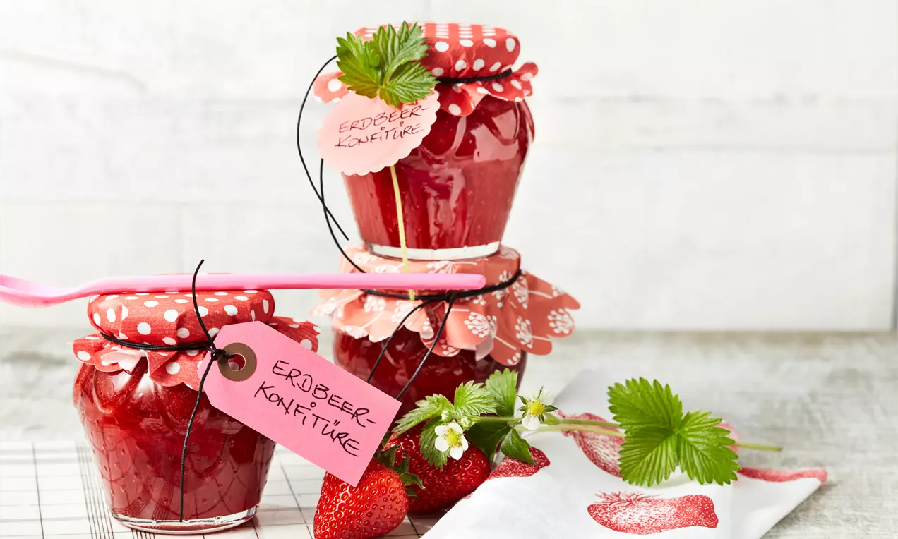 Erdbeerrote Konfitüre in zwei gläsernen Schraubgläsern mit gepunktetem Stoffdeckel, dekoriert mit frischen Erdbeeren und grünen Blättern