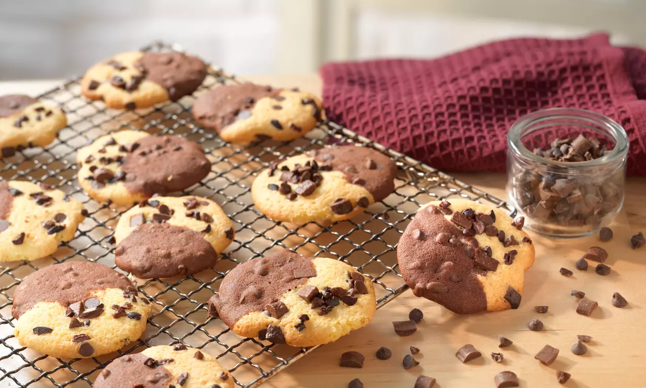 Schwarz-Weiß-Cookies mit weicher Textur halb dunkelbraun halb goldgelb, bestreut mit Schokostückchen auf einem Metallgitter neben rotem Geschirrtuch