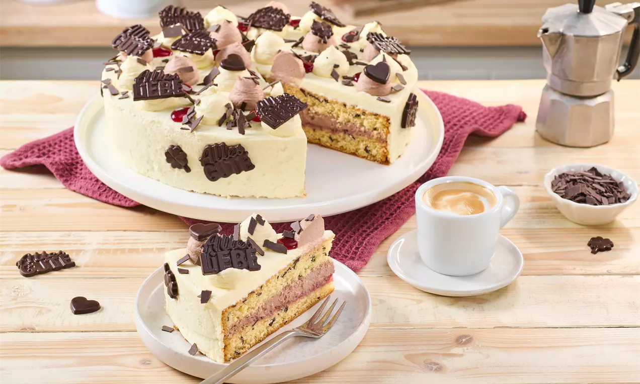 Geburtstagstorte mit heller Creme und Schokodekor auf einer weißen Platte, gefüllt mit einer rosa Schicht. Ein Stück liegt auf einem Teller mit Gabel, daneben eine Tasse Espresso und Schokostückchen.