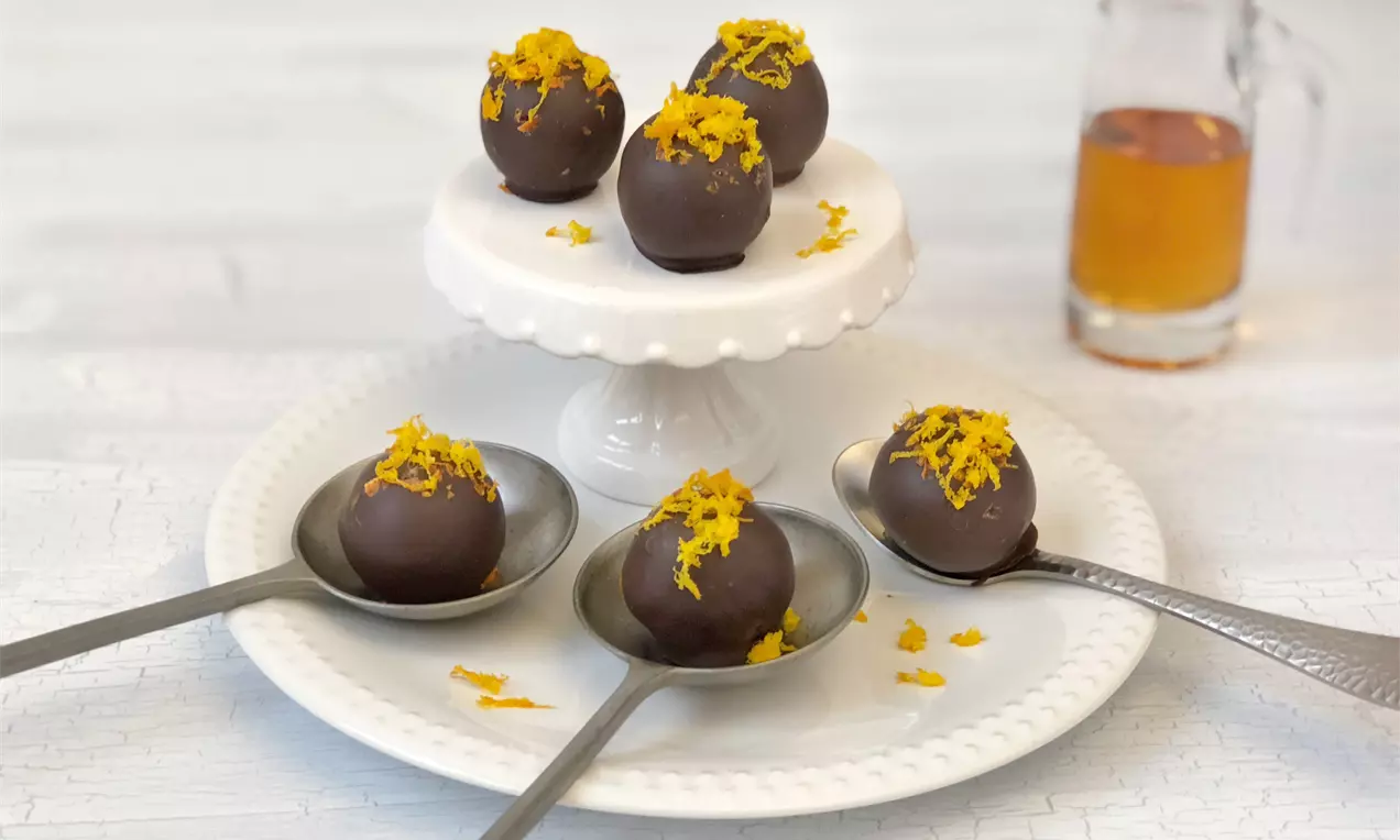Kugeln aus glänzend dunkler Schokolade mit leuchtend orangefarbenen Zesten, dekorativ auf silbernen Löffeln und einer weißen Etagere arrangiert
