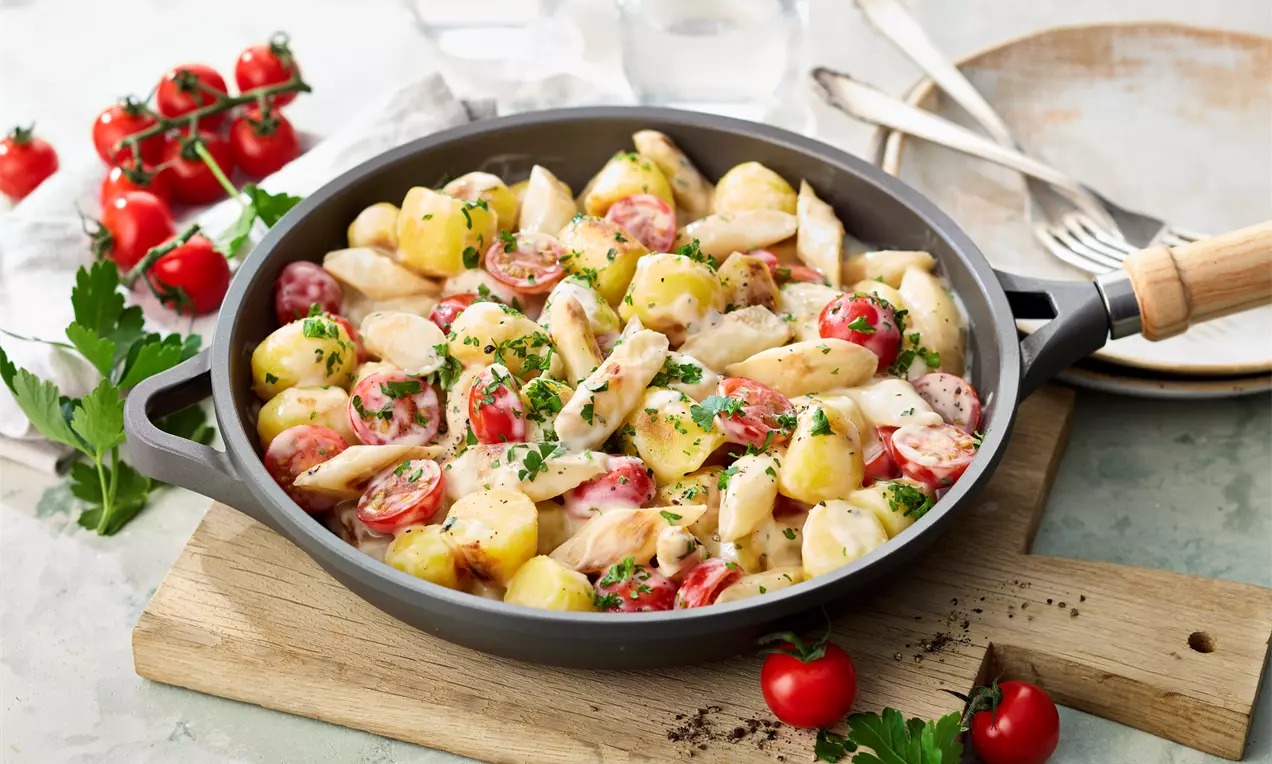Bissfeste Spargelstücke, goldgelbe Kartoffelwürfel und saftige Kirschtomaten in cremiger Sauce, bestreut mit Kräutern in grauer Pfanne