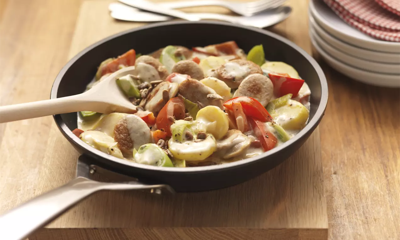 Bratwurstscheiben mit goldbraunem Rand, Paprika, Champignons und Frühlingszwiebeln in cremiger Soße serviert in schwarzer Pfanne