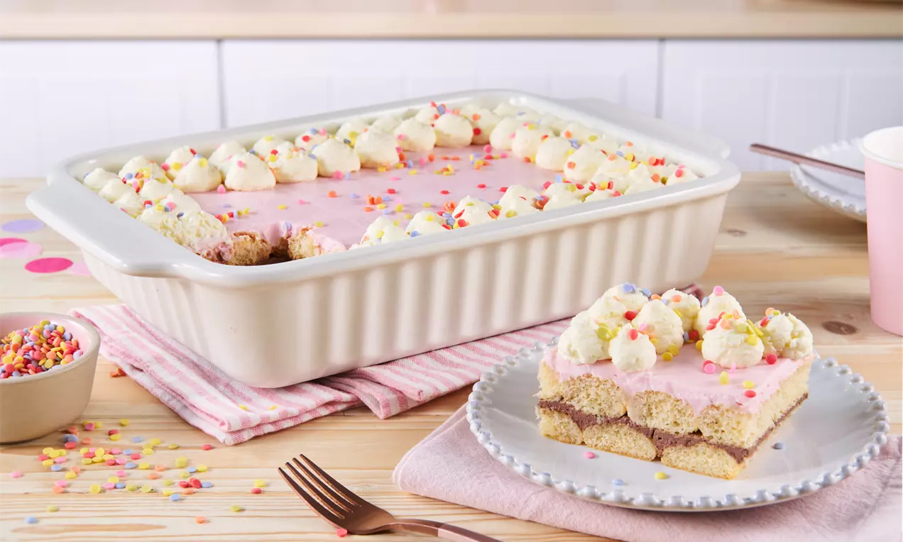 Eine rechteckige Torte mit rosa Glasur, bunten Zuckerstreuseln und Sahnetupfen in einer hellen Backform auf einem Holztisch mit rosafarbenen Details