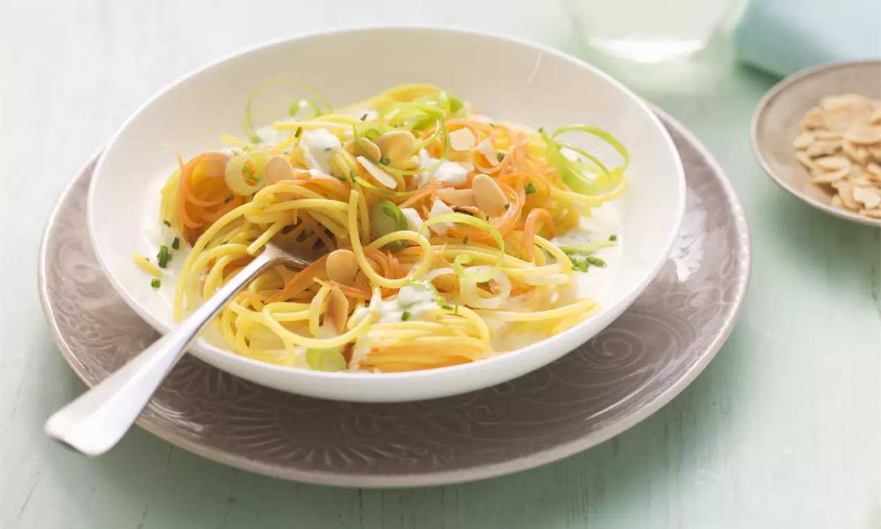 Spaghetti mit gelben und orangenen Möhrenstreifen, garniert mit Mandelblättchen und Lauchzwiebeln auf weißem Teller mit grauem Unterteller
