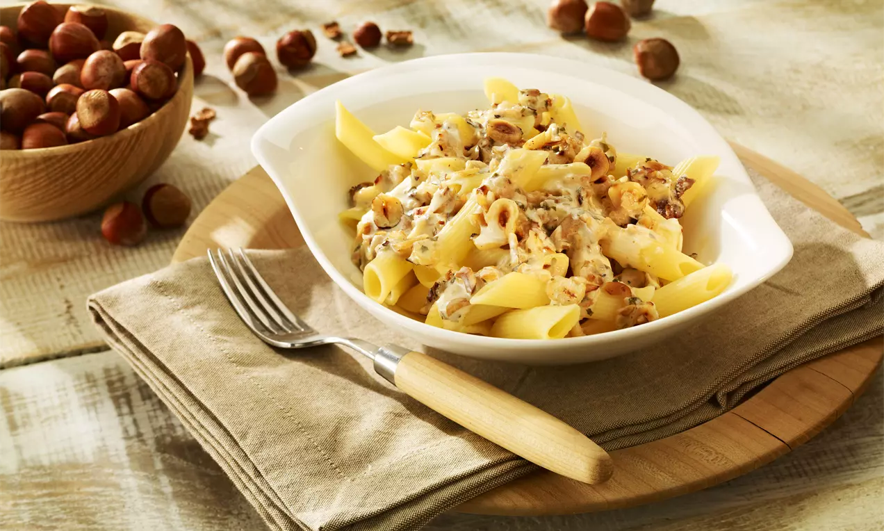 Penne-Nudeln in cremiger beige-brauner Haselnuss-Soße, garniert mit gehackten Haselnüssen, serviert in einer weißen Schale auf Holzbrett und Stoffserviette