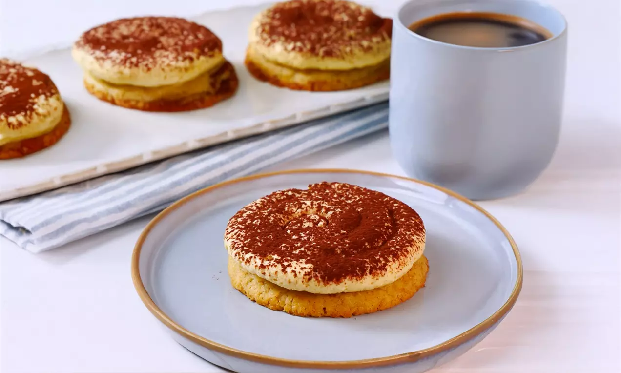 Tiramisu-Crumbl-Cookies auf Tellern und einem Tablett, mit kakaobestäubter Creme und einer Tasse Kaffee daneben auf einem hellen Untergrund.