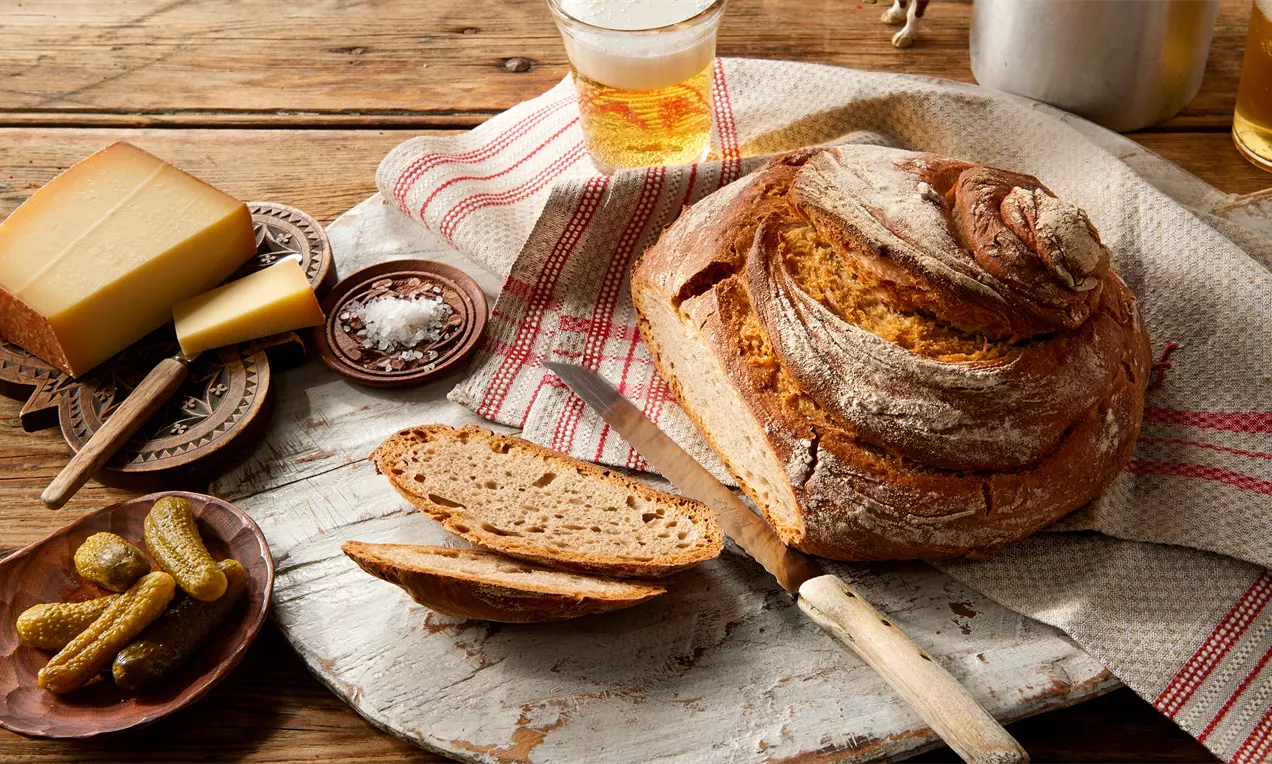 Rustikales St. Galler Landbrot mit goldbrauner Kruste und luftiger Textur, begleitet von Käse, Gurken und Salz auf Holzbrett und Stofftuch