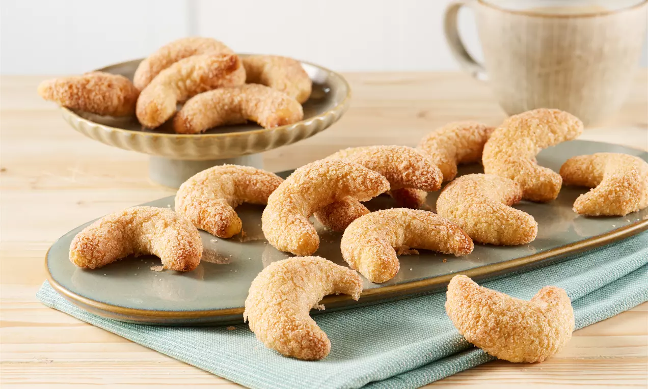 Churro-Kipferl in goldbrauner Zimtzuckerkruste, halbmondförmig angeordnet auf einer rechteckigen Platte mit hellblauem Stoffuntergrund