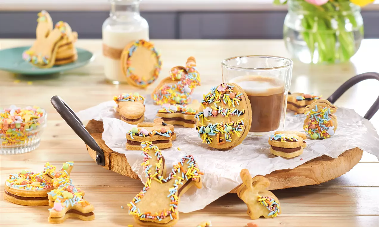 Gefüllte Osterkekse in Hasenform mit goldbrauner Kruste und dunkler Nougatfüllung, halbiert auf Teller und mit bunten Streuseln bestreut