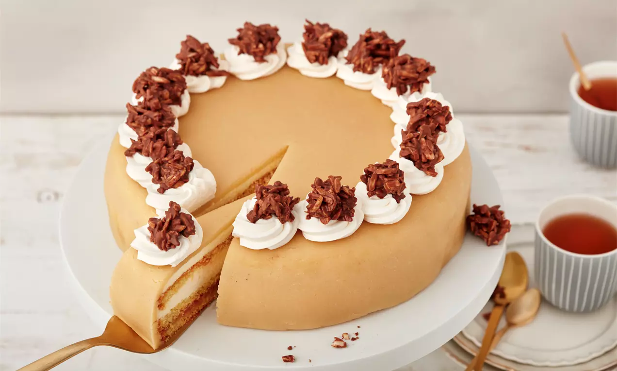 Lübecker Marzipantorte mit hellbrauner Marzipandecke, Sahnetupfen und Schokoladenornamenten auf weißer Tortenplatte mit angeschnittener Stück