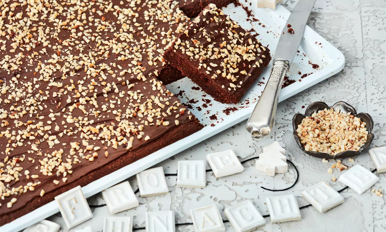 Schokoladenbrot mit glänzender Schokoladenglasur und gehackten Nüssen, auf einem weißen Teller mit Dekoration und Messer angerichtet