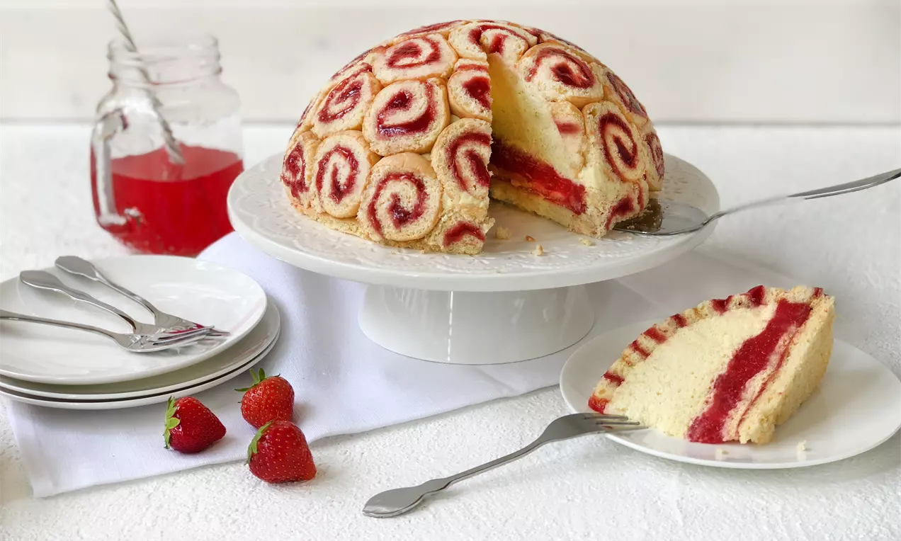 Halbkugelförmige Erdbeer-Charlotte mit spiralförmigen Biskuitrollen, roter Fruchtfüllung und heller Creme auf weißer Tortenplatte angerichtet