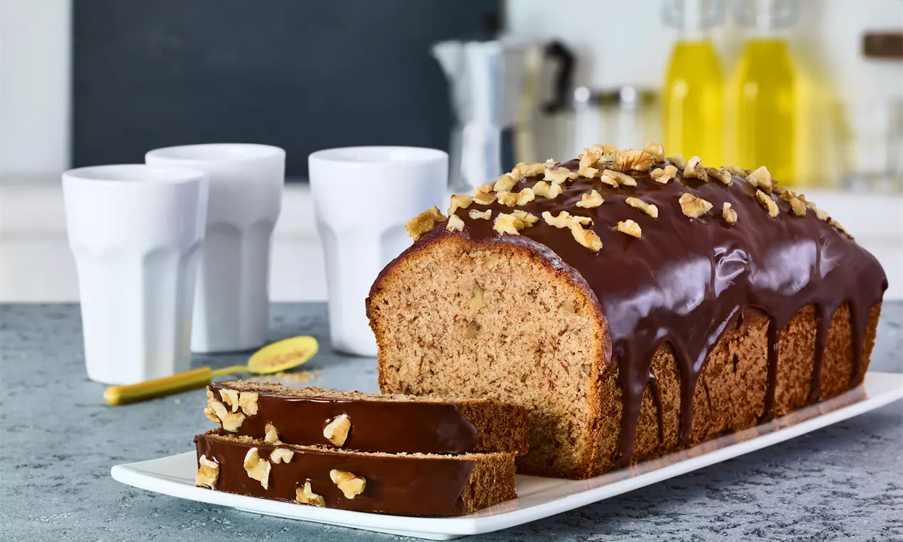 Saftiges Banana Bread mit Walnüssen und glänzendem Schokoladenguss auf einer weißen Platte, daneben drei Gläser auf grauem Untergrund