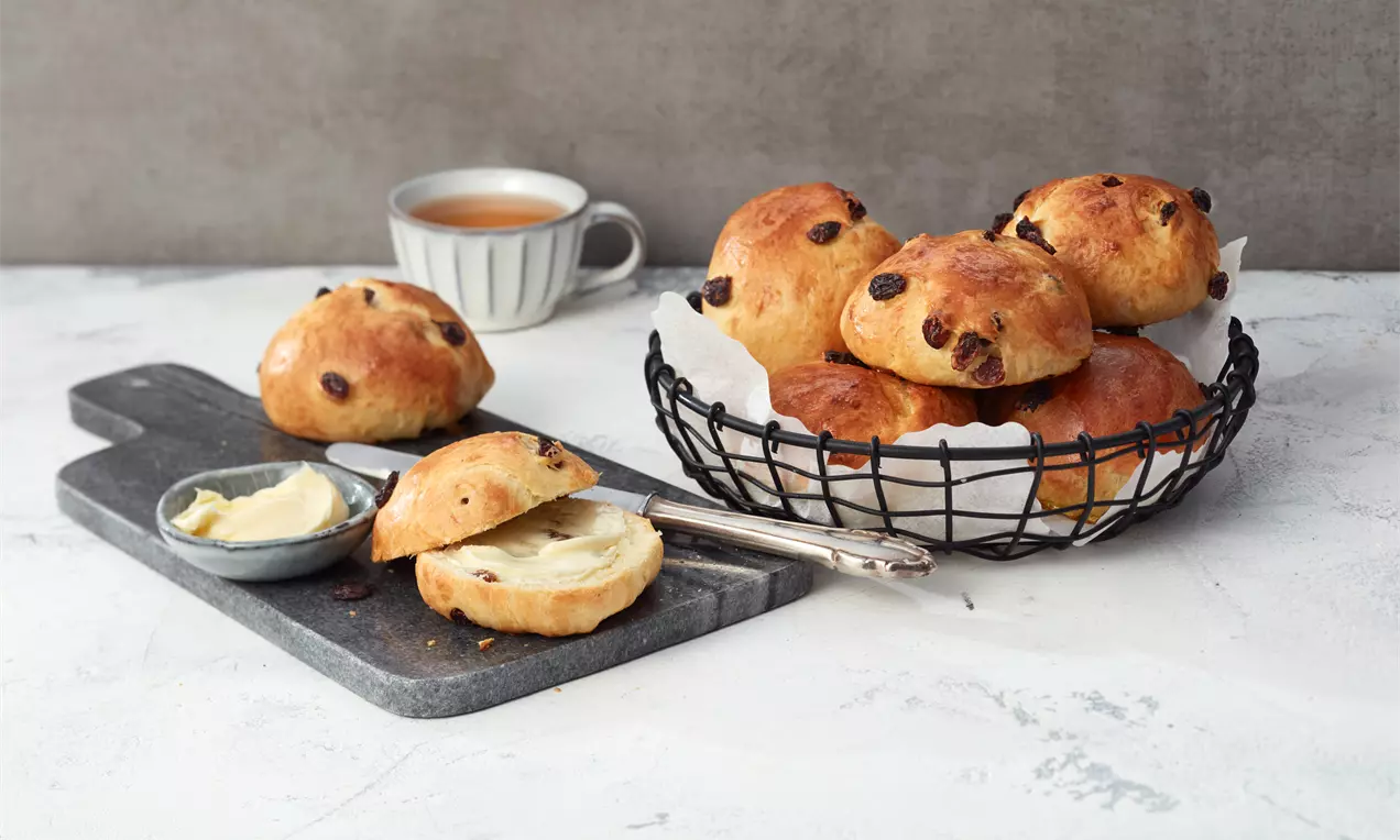 Runde, goldbraune Rosinenbrötchen mit glänzender Oberfläche in einem schwarzen Korb, dazu ein aufgeschnittenes Brötchen mit Buttercreme auf einem grauen Brett