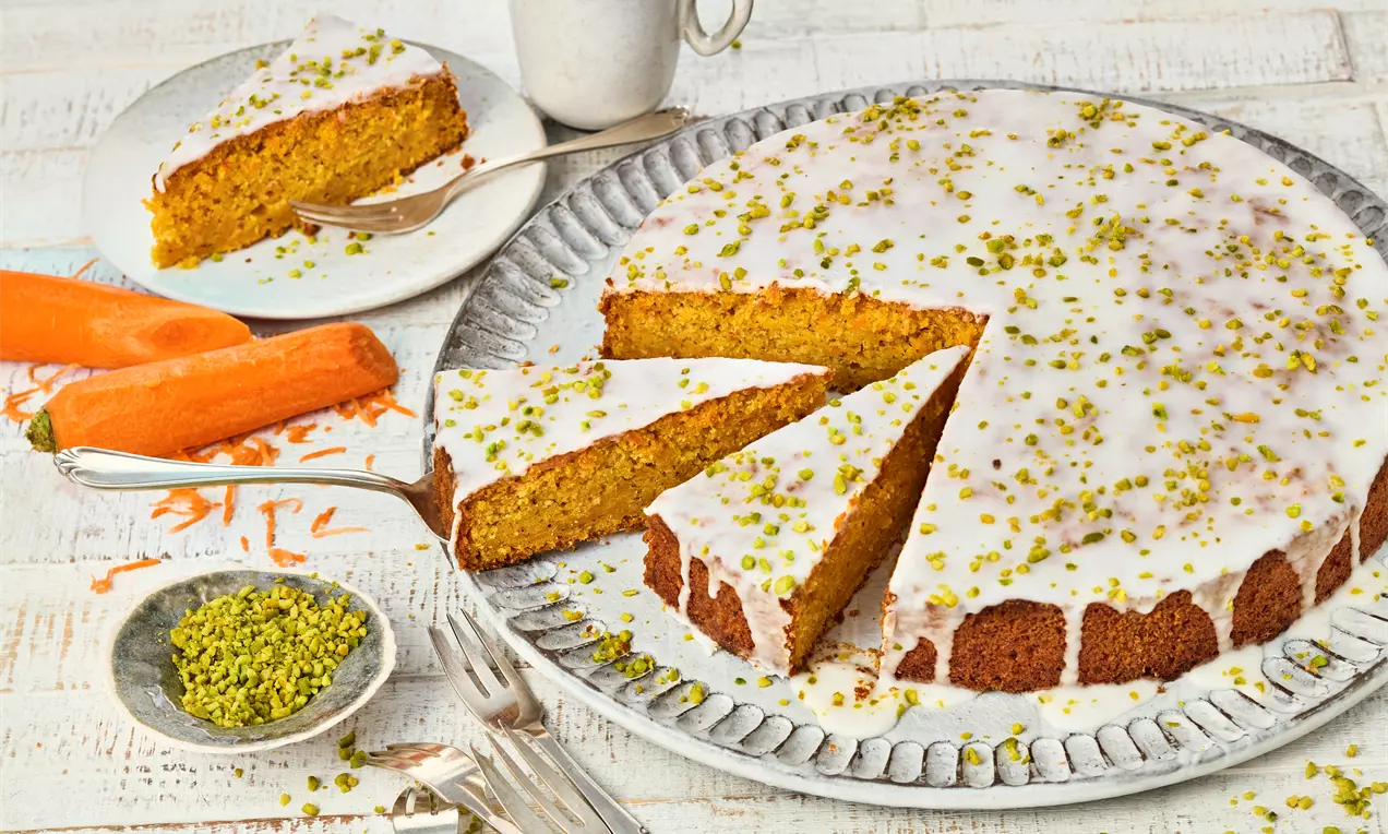Veganer Karottenkuchen auf einer Platte, mit weißer Glasur und gehackten Pistazien bestreut. Ein Stück ist herausgeschnitten. Daneben Karotten, Pistazien, Besteck und eine Tasse Kaffee.