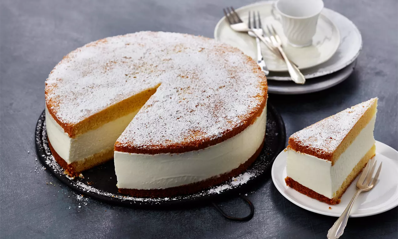 Käse-Sahne-Torte mit goldbraunem Biskuitboden und -deckel, weißer Sahnefüllung und Puderzucker, auf einer schwarzen Servierplatte mit Teller und Gabel