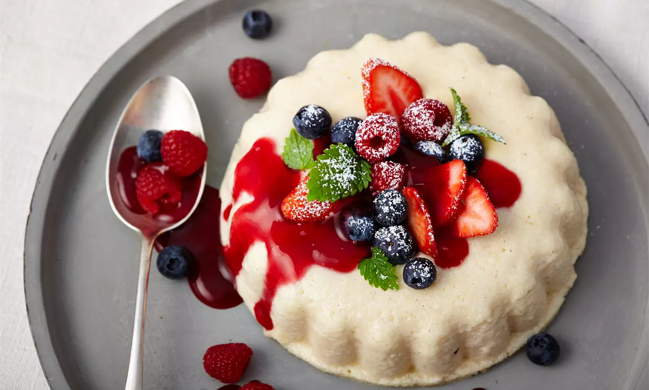 Grießpudding in Gugelhupfform auf grauem Teller, garniert mit roten Beeren, Blaubeeren, Minzblättern, Puderzucker und roter Fruchtsauce