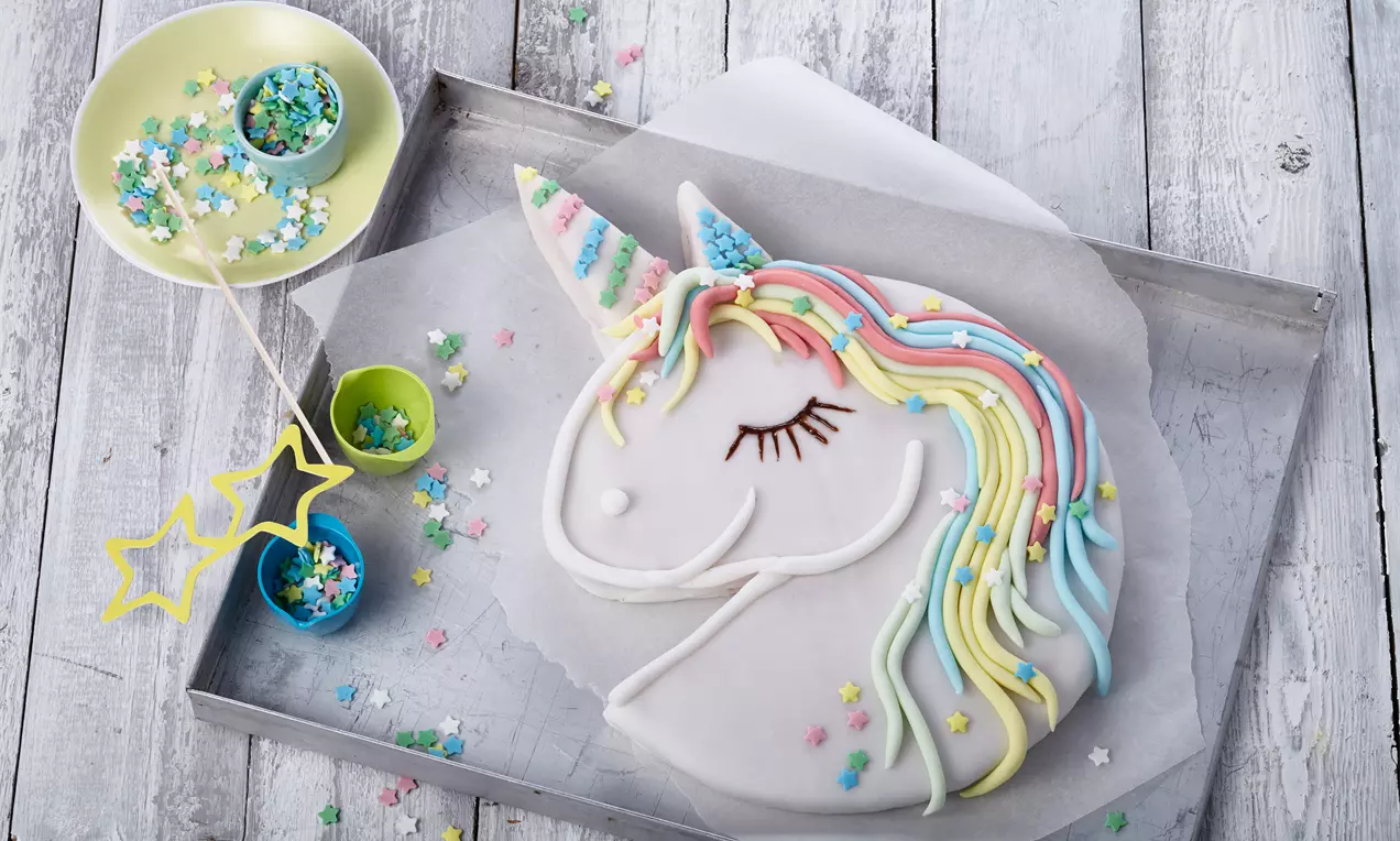 Einhörnerförmiger Kuchen mit weißem Fondant, bunten Zuckerglasur-Mähnen, pastellfarbenem Horn und Dekor auf heller Holzplatte