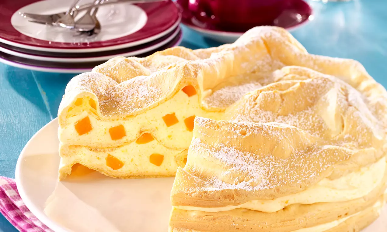 Brandteigtorte mit goldgelbem, fluffigem Teigdeckel, Aprikosenstückchen in hellgelber Cremefüllung, leicht mit Puderzucker bestäubt auf weißem Teller