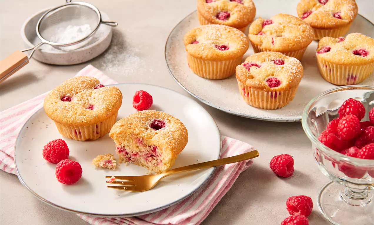 Saftige Himbeer-Muffins mit goldgelber, lockerer Kruste und roten Himbeeren, angerichtet auf Tellern mit frischen Beeren und dekorativem Sieb