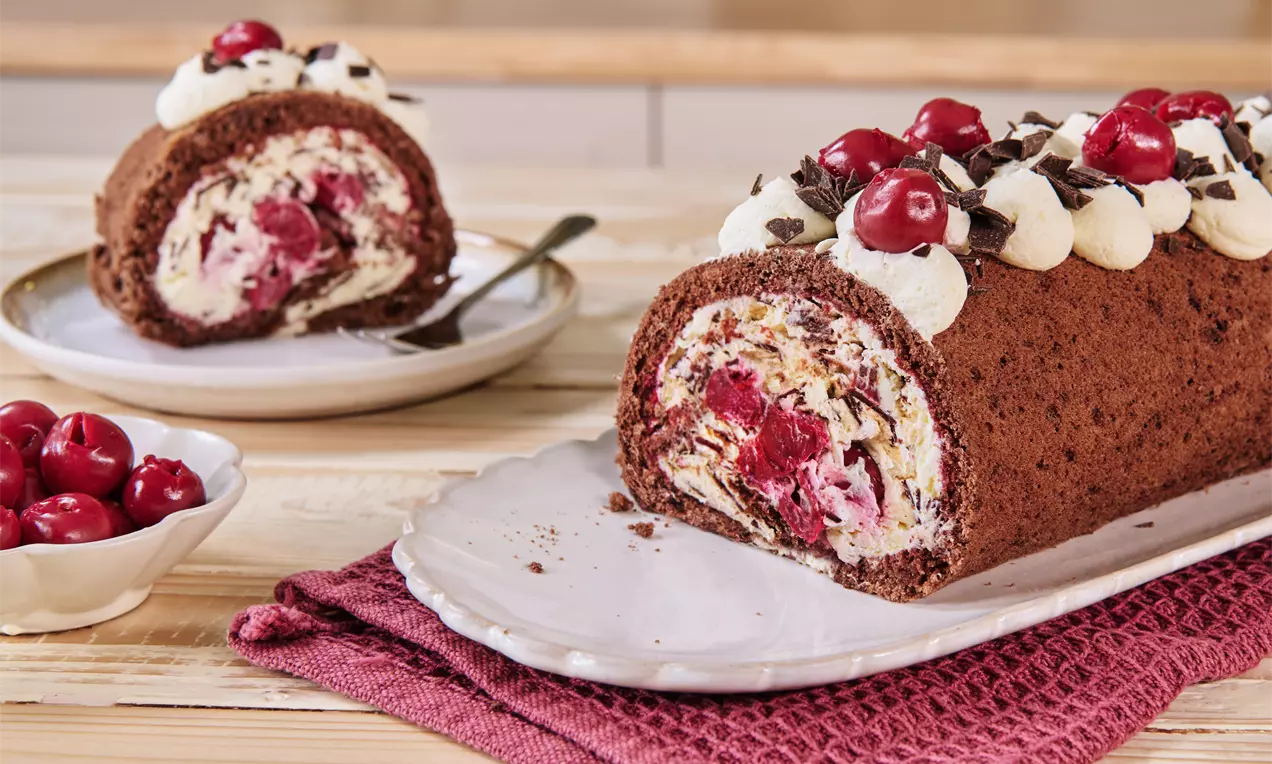 Schokoladen-Biskuitrolle mit Sahnecreme, saftigen Kirschen und Schokoraspeln, auf weißer Platte mit roten Kirschen in Schale angerichtet