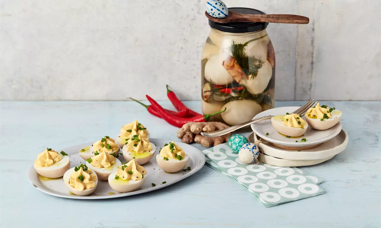 Halbierte Soleier mit cremig-gelber Füllung, bestreut mit feinen grünen Kräutern, auf weißer Platte und Tellern neben Einmachglas und Deko