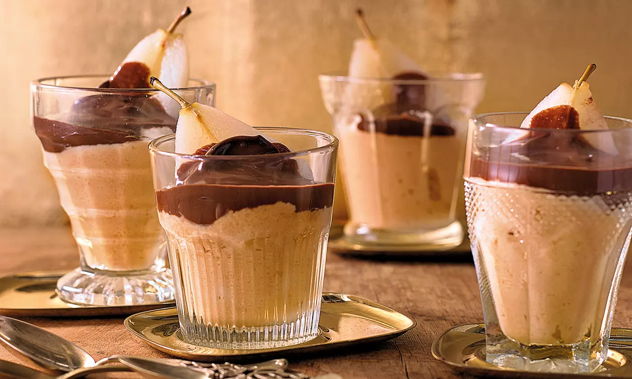 Karamellfarbene Mousse in Gläsern mit glatter Schokosoße und halbierten Birnen garniert auf goldenen Tellern vor einem goldenen Hintergrund