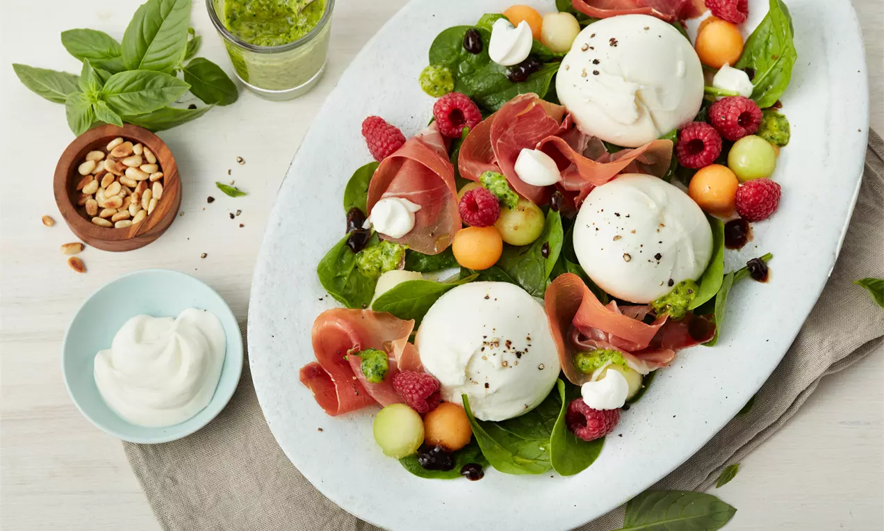 Burrata mit Melone, Parma-Schinken und Basilikum-Pesto, cremig weiß, leuchtend orange Melonenstücke, rosa Schinken und grüne Basilikumblätter