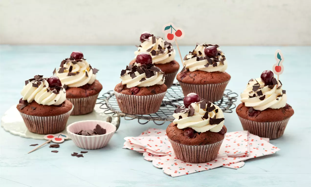 Schwarzwälder Kirsch-Cupcakes aus saftigem Schokoladenteig mit Sahnetopping, Kirschen und Schokospänen auf einem hellblauen Tisch angerichtet