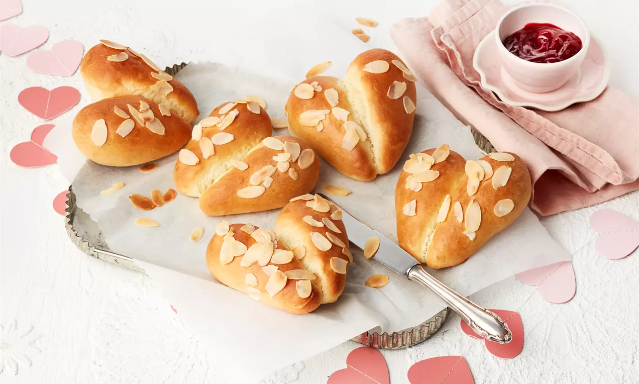 Goldbraune, herzförmige Hefebrötchen mit Mandelblättchen auf Backpapier, daneben rote Konfitüre in einer rosa Schale und rosa Deko-Herzen