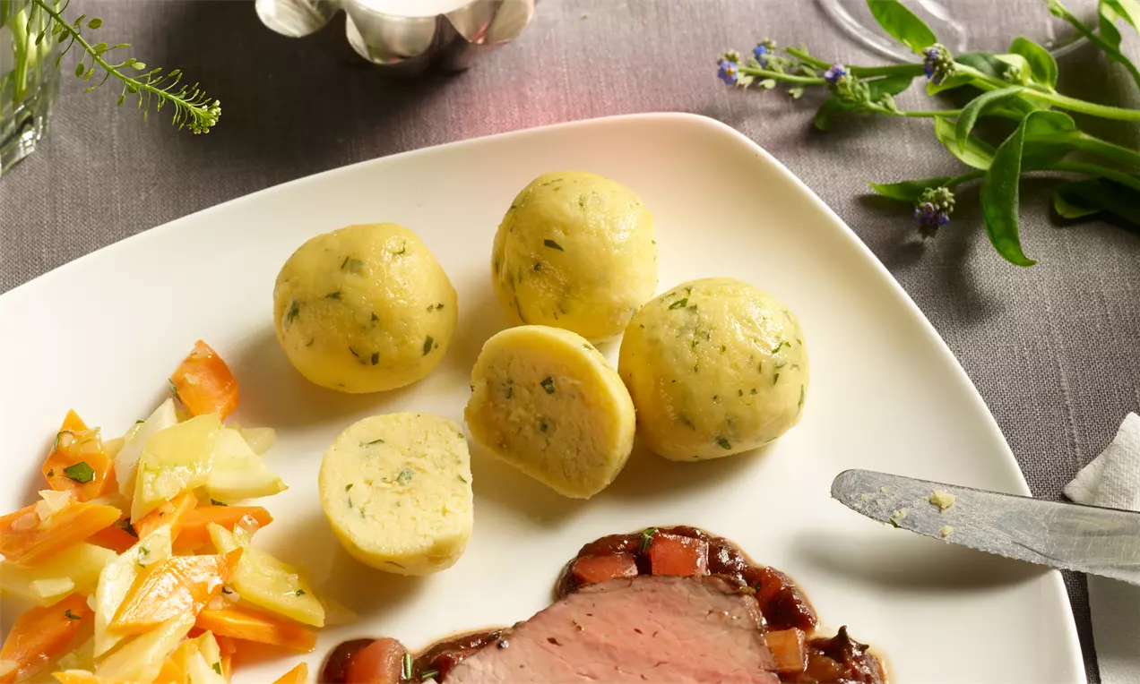 Grießknödel, rund und hellgelb mit grünen Kräuterpunkten, weich texturiert, neben Gemüse und Braten auf einem weißen Teller angerichtet