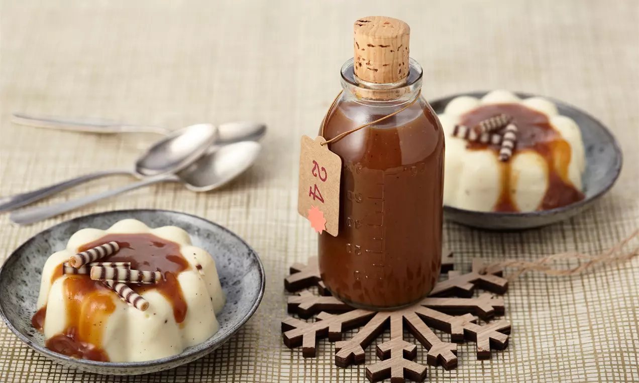 Glasflasche mit brauner Toffeesoße auf Holzschneeflocke, daneben Panna-Cotta in grauen Schalen mit Karamellsoße und Schokoröllchen
