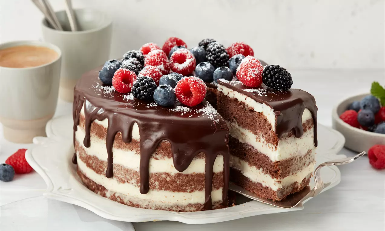 Drip-Cake mit Schichten aus weißer Creme und braunem Biskuit, überzogen mit dunkler Schokolade, garniert mit Beeren und Puderzucker auf einer Tortenplatte