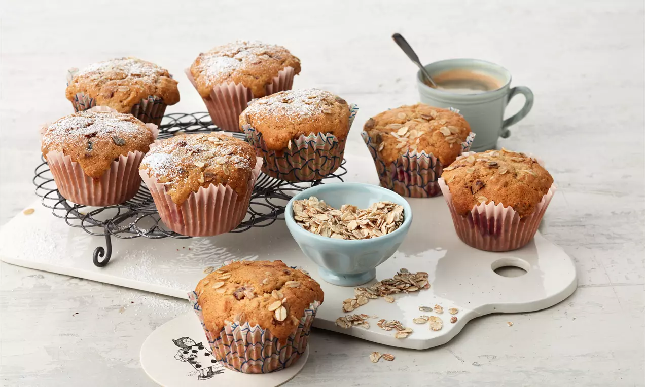 Goldbraune Müsli-Muffins mit Haferflocken bestreut in Papierförmchen auf einem weißen Brett und einer Metallplatte neben einer Tasse Kaffee