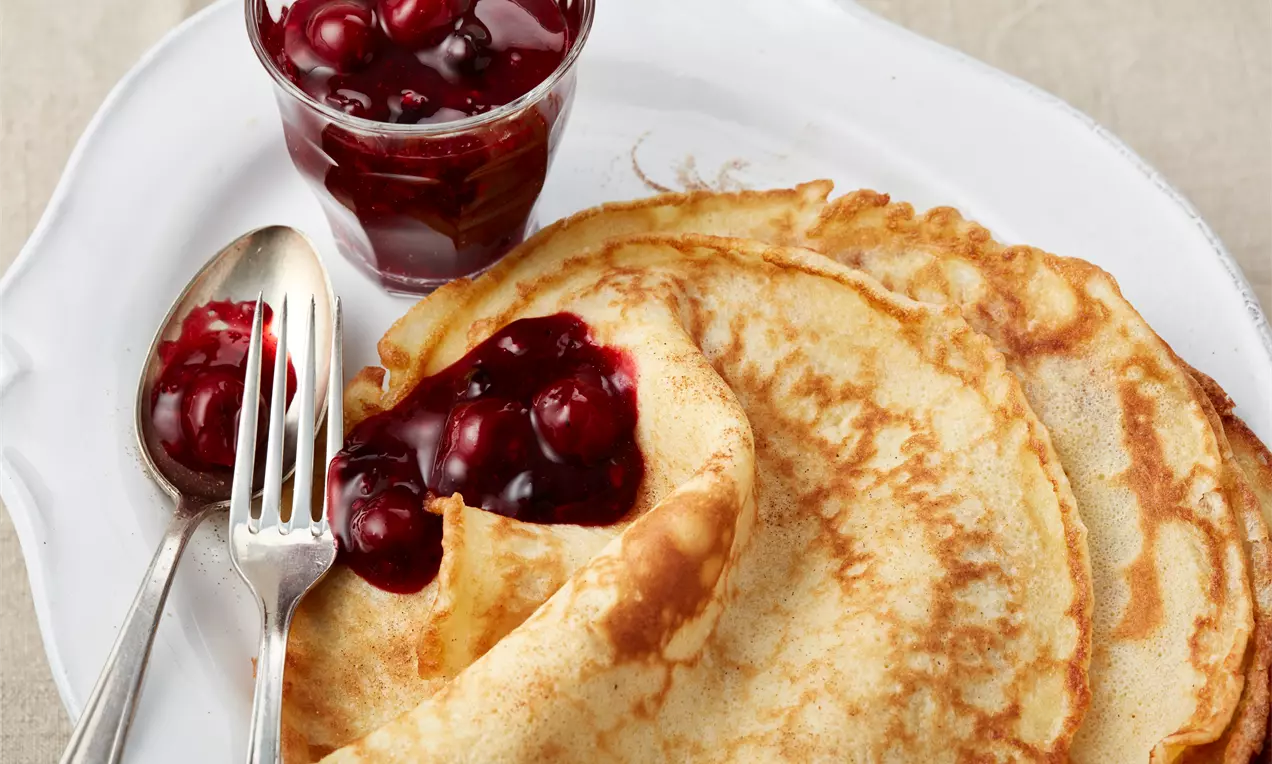 Dünne, goldbraune Pfannkuchen mit leichter Bräunung, angerichtet auf weißem Teller, dazu ein Glas rote Kirschfüllung und Besteck