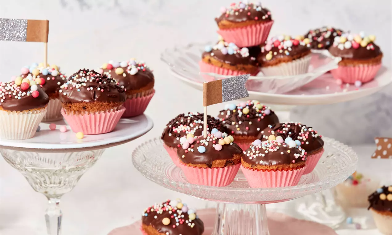 Mini-Muffins mit glänzendem Schokoladenguss, bunten Zuckerstreuseln, in rosa Förmchen auf gläsernen Etageren arrangiert