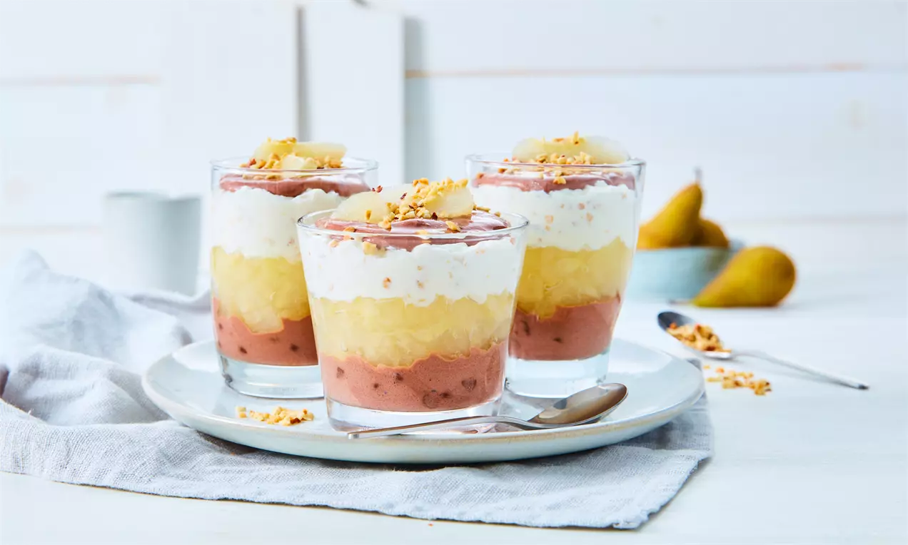 Schichtdesserts in Gläsern mit roten, gelben und weißen Lagen aus Frucht und Creme, garniert mit Nüssen, auf hellem Teller angerichtet