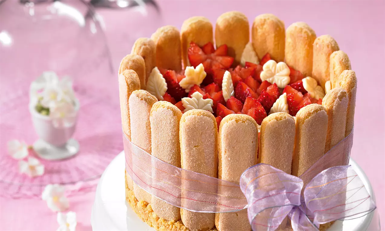 Goldbraune Löffelbiskuits, zu einem Kranz geformt und mit rosa Schleife gebunden, umgeben einen Kern aus Erdbeerstückchen und Sahnetupfen