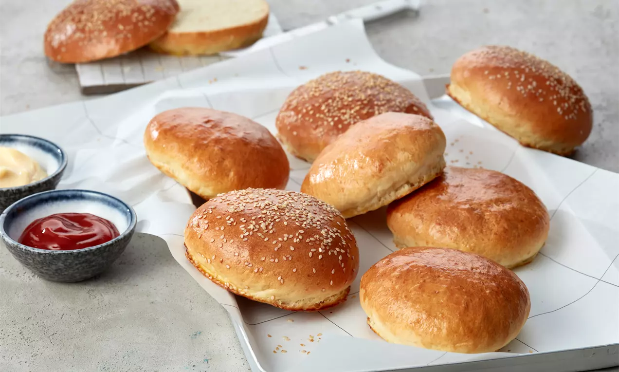Goldbraune Burger-Brötchen mit glatter Oberfläche und Sesam bestreut, locker arrangiert auf einem weißen Tablett neben Schalen mit Ketchup und Mayonnaise