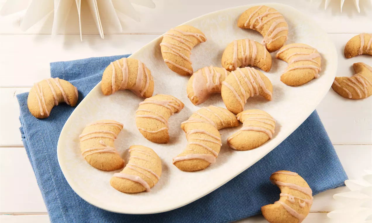 Goldbraune, geschwungene Lebkuchen-Kipferl mit glatter Glasur auf einem ovalen Teller, angerichtet auf einem blauen Stofftuch
